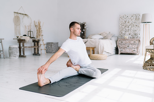 Pensive calm male in casual wear enjoying morning yoga zen feeling body and stretching muscles, handsome hipster guy keeping healthy lifestyle resting during yoga training at apartment on carpet