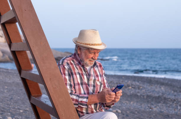 istock Smiling caucasian senior man with hat sitting on the beach at sea while using mobile phone, bearded male enjoying free time, vacation or retirement 1425212535