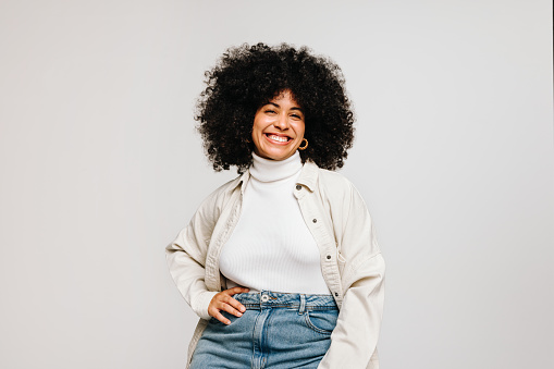Confident young woman with an Afro hairstyle smiling at the camera while standing against a white background. Happy woman of colour wearing her natural curly hair with pride.