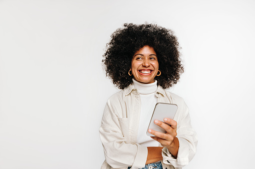 Happy young woman looking away with a smile while holding a smartphone in her hand. Beautiful woman with curly hair contemplating an idea while standing against a studio background.