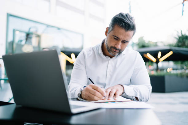 istock Mature entrepreneur writing informative notes in textbook sitting in sidewalk cafeteria with modern netbook and working remotely, middle aged proud CEO planning business meeting in financial district 1425218539