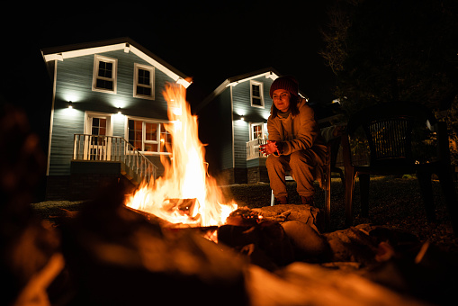 Backyard of a wooden house at night. A woman covered with a plaid sits by the fire.