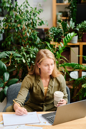 Young creative woman with long blond hair having coffee and making notes in document while analyzing online data on laptop screen