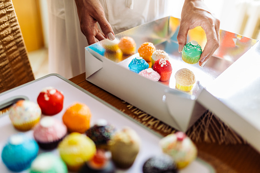 Young woman preparing home made cakes for delivery. She is packing colorful cookies in a delivery box. She is wearing white bathrobe and working in her kitchen. Online shopping and subscription services concept