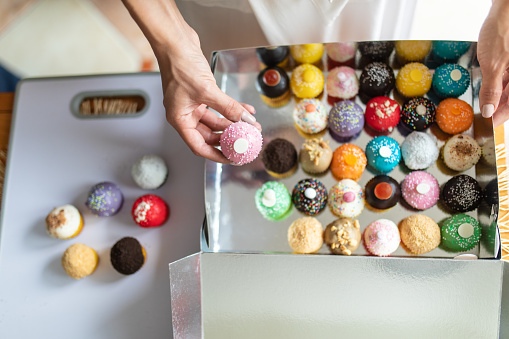 Young woman preparing home made cakes for delivery. She is packing colorful cookies in a delivery box. She is wearing white bathrobe and working in her kitchen. Online shopping and subscription services concept