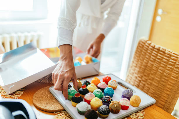 Young woman preparing home made cakes for delivery. She is packing colorful cookies in a delivery box. She is wearing white bathrobe and working in her kitchen. Online shopping and subscription services concept