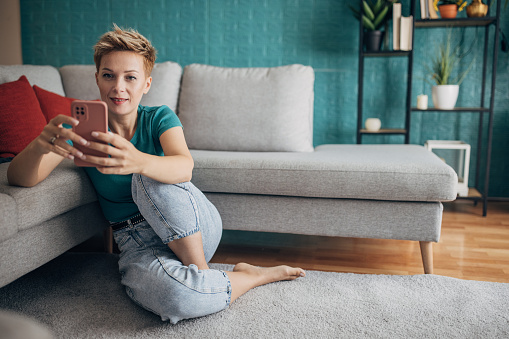 Beautiful shorthaired woman sitting on the floor in living room at home, she is using a smart phone.