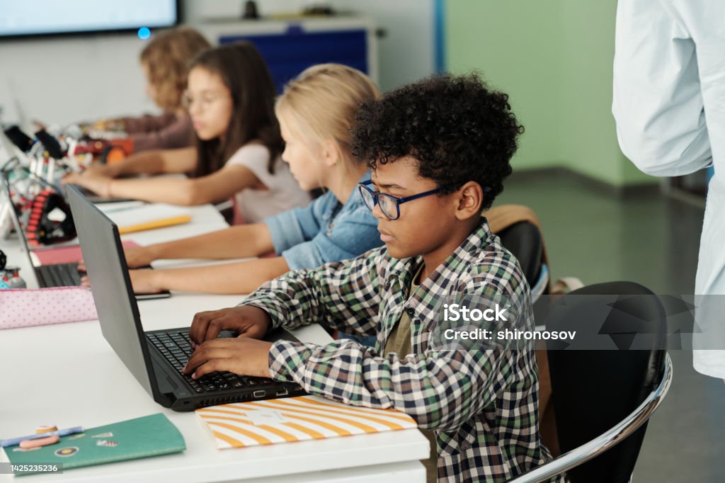 Side view of youthful African American schoolboy working in front of laptop Side view of youthful African American schoolboy and his classmates working in front of laptops while sitting in row by desk Child Stock Photo