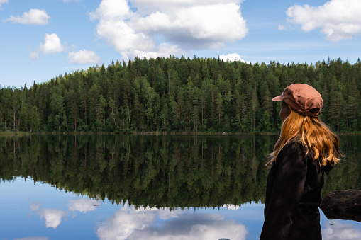 Red-haired woman wearing a cap looking at the calm lake with reflection on a beautiful summer day in nature