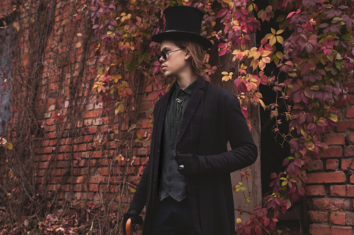 Gothic man with long hair in glasses and hat. Black classic clothes. Vampire in black suit on autumn background. Halloween costume. Portrait