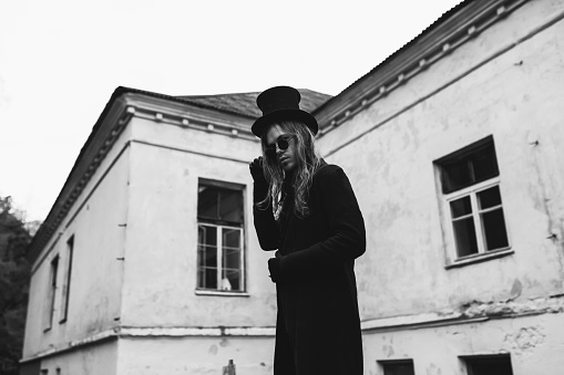 Gothic man with long hair in glasses and hat. Black classic clothes. Vampire in suit on autumn background. Halloween costume. Portrait. Black and white photography