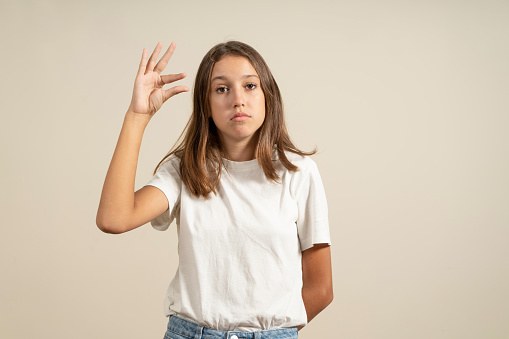 Portrait of cute teen girl in white t-shirt showing small size or small gesture with fingers, looking up with imploring grin. Indoor studio shot isolated on beige background