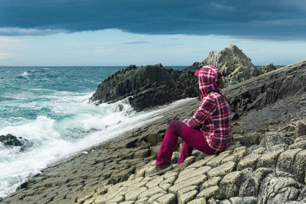 traveler sits on the shore of a stormy sea, on a natural pavement made of columnar granite traveler sits on the shore of a stormy sea, on a natural pavement made of columnar granite kunashir island stock pictures, royalty-free photos & images