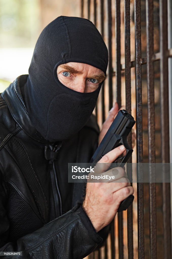 Scary man masked by balaclava and holding a gun approaches the barred window of a house Ominous mature man wearing ski mask about to carry out a murder or assassination. 50-59 Years Stock Photo