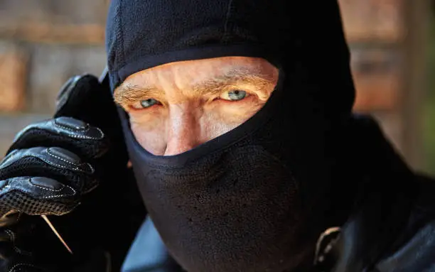 Ominous mature man wearing ski mask holds a cellphone to his ear.