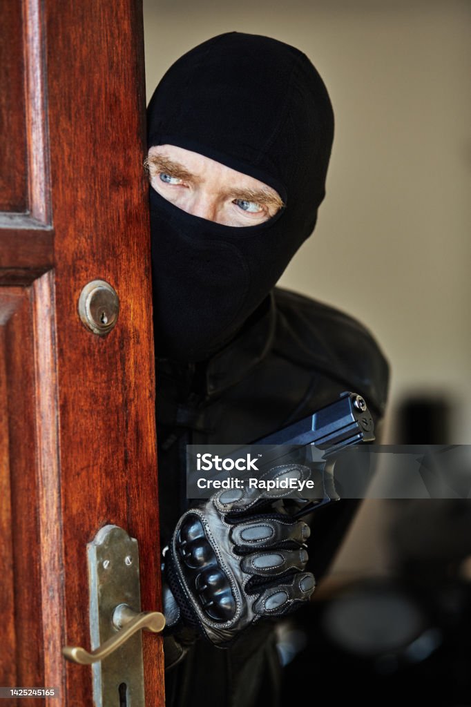 Scary man masked by balaclava looks furtively around a door, holding a gun in his gloved hand Ominous mature man wearing ski mask and holding a gun, giving a suspicious sideways glance. Doorway Stock Photo