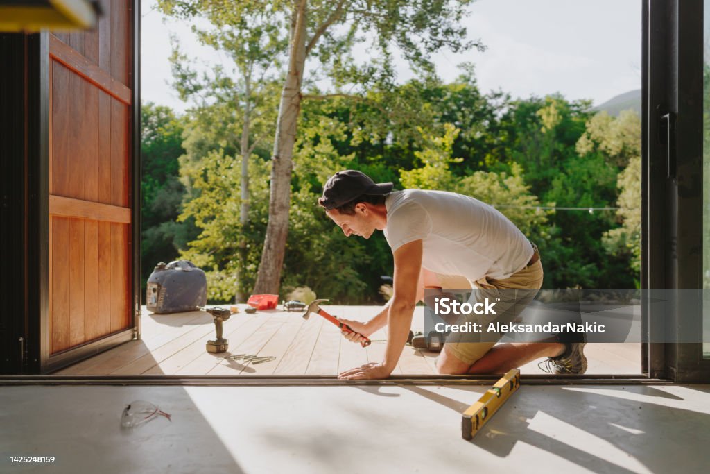 Man working on wooden decking in front of a cabin house Photo of a carpenter installing wooden decking in front of a cabin house Deck Stock Photo