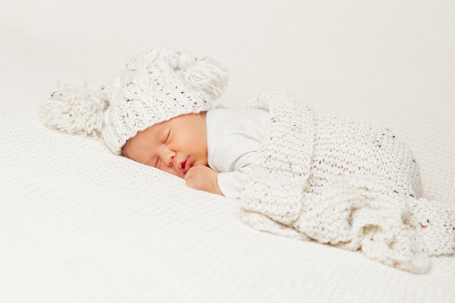 Baby sleeping with Blanket. Newborn lying on Stomach in White Bed. Cute Baby Face Portrait in Knitted Hat. One Month Child Bedtime