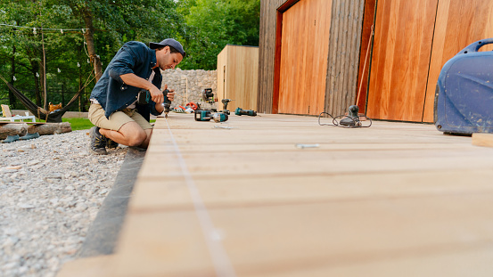 Photo of a carpenter installing wooden decking in front of a cabin house