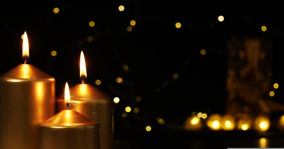 The lights of a Christmas garland glow mysteriously in the background of three burning golden candles. The flame of the candle burns steadily. Concept for Christmas cards and backgrounds.