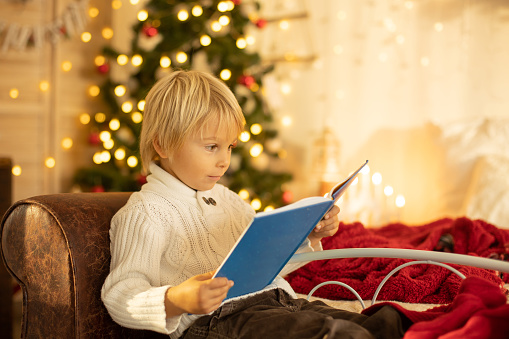 Cute blond toddler preschool boy, reading a book and opening present on Christmas on cozy home, lights and decoration