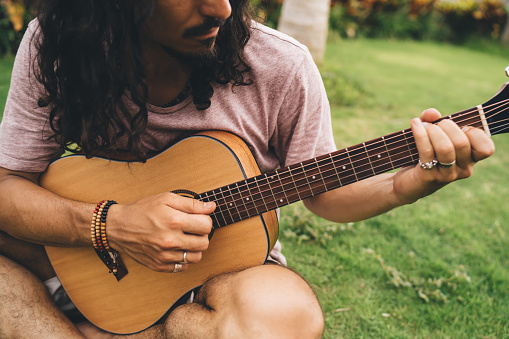 Crop ethnic man in casual wear with long black hair and mustache sitting on green grass and chill while playing guitar