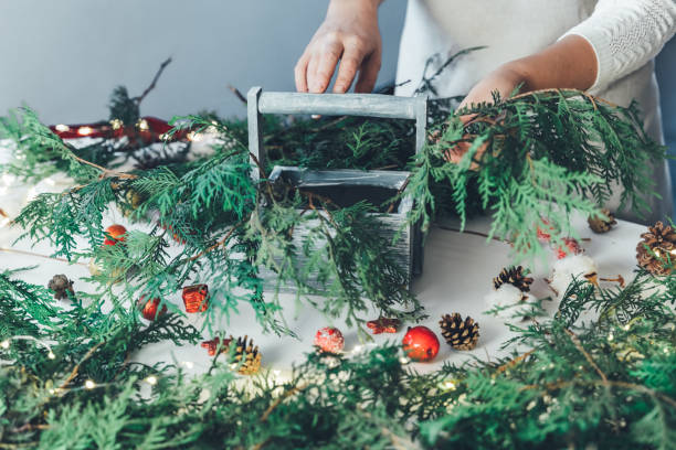 istock Florist woman makes Christmas composition for table decoration from thuja, spruce branches in wooden box. Using of floral foam, cones, snow apples, balls, cotton flowers,garland. New Year celebration 1425260148