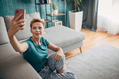 Beautiful shorthaired woman sitting on the floor at home, she is using a smart phone to take a selfie.