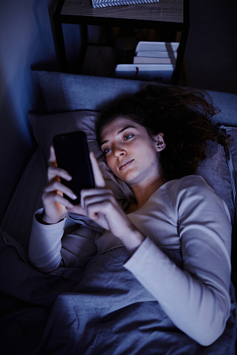High angle view of young woman communicating online on mobile phone before sleep lying on her bed