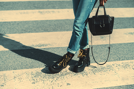 Close-up view of legs of black woman, in ripped jeans and with black glossy leather boots with yellow laces, standing on the zebra of a pedestrian crosswalk on the road with an elegant bag in the hand