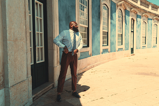 A fashionable mature bearded bald black businessman in a white shirt, glasses, and brown trousers is adjusting his belt while standing next to the door of an antique building with stained windows