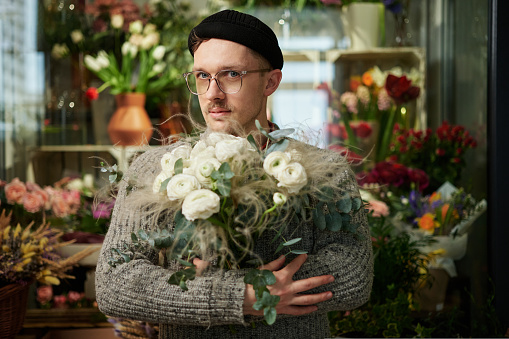 Caucasian man in grey sweater, beanie hat and eyeglasses holding beautiful bouquet of white fresh rose flowers. Love, dating, romance, Mother's Day or Valentine's Day concept. Congratulating concept