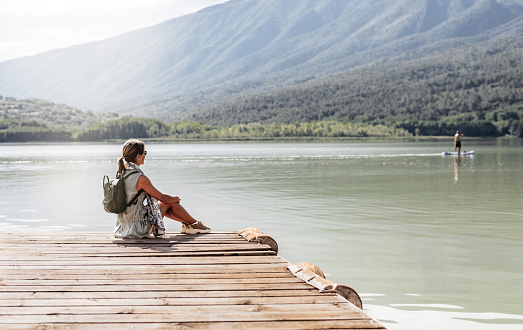 General side view of a woman with a backpack sitting on a wooden dock of a lake, looking at a person paddle surfing. concept of tranquility and relaxation.