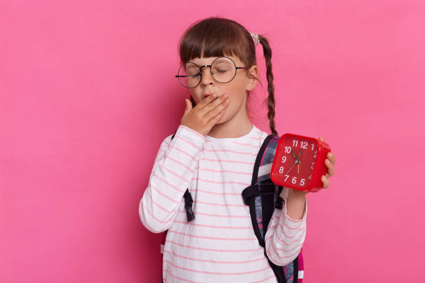 셔츠와 안경을 쓰고 분홍색 배경에 고립 된 배낭으로 서서 빨간 알람 시계를 들고 하품을하고, 손바닥으로 입을 가리는 졸린 피곤한 어린 여학생의 수평 촬영. - classroom bell child elementary school 뉴스 사진 이미지