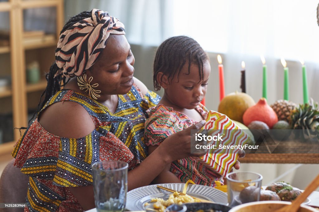Mom opening gift with her child African woman in national costume opening present with her little daughter while they sitting at dining table Kwanzaa Stock Photo