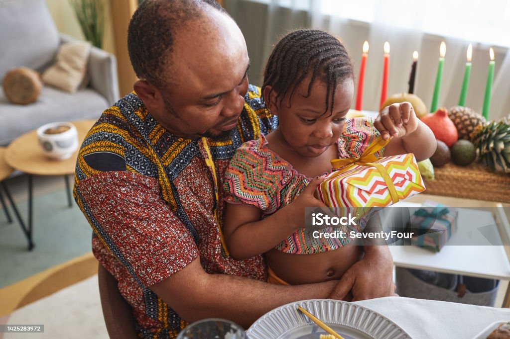 Little girl opening gift box African little girl opening gift box while sitting on knees of her father at dining table Kwanzaa Stock Photo