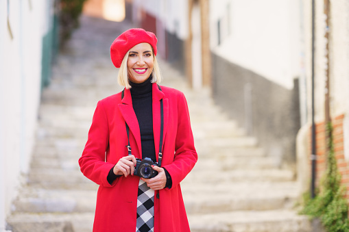 Confident young positive woman, with blond hair in fashionable red coat and beret smiling and looking away while standing on stone stairs with photo camera during trip in old town