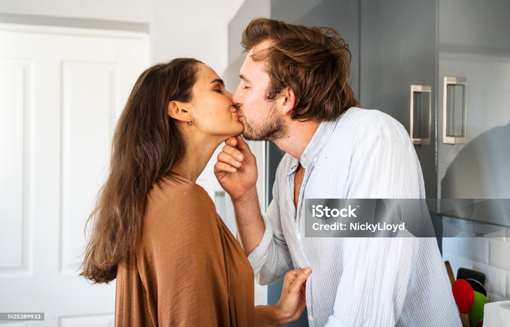 Young woman kissing her boyfriend in their kitchen Affectionate young woman kissing her boyfriend sitting on a counter in their kitchen at home Couple - Relationship Stock Photo