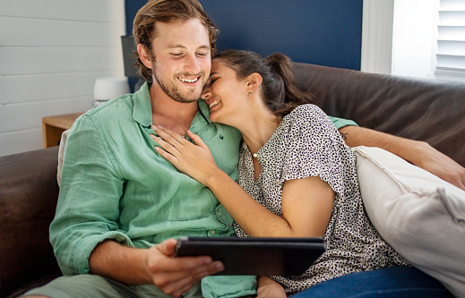Laughing young couple streaming a video on a digital tablet while relaxing on their living room sofa