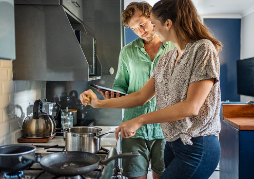 Smiling young couple using an online recipe on a digital tablet to make dinner together on their kitchen stove