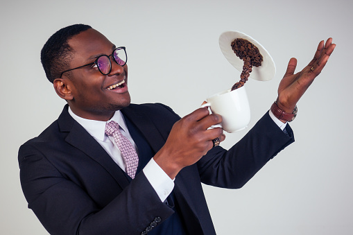 surprised and amazed handsome afro american business man in a black classic suit and glasses holding a cup with flying coffee bean splash on plate in white background studio shot. Magic morning drink