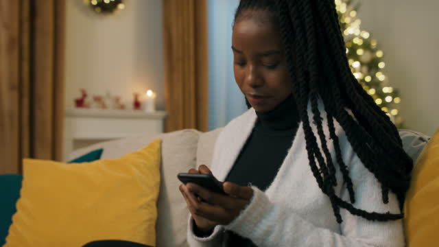 Attractive girl of African appearance sitting on the couch and holding phone. Girl looking for Christmas decor for home. She looks at decorations and chooses what she likes