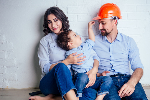 happy family handsome man in helmet hardhat and charming woman and their cute little son sitting on the floor against a white wall . concept of buying repairs house apartment by young family.