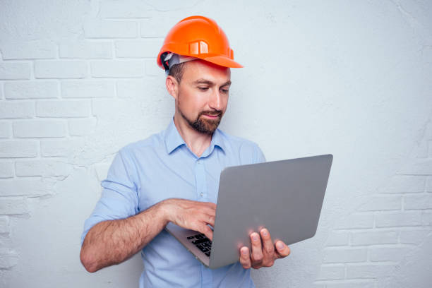 istock handsome man with a beard and a helmet hardhat on a background of a white brick wall builder holding a laptop online purchase of building materials 1425297530