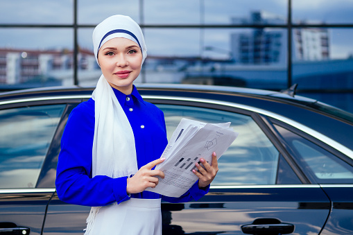 beautiful and successful european muslim business woman entrepreneur in stylish hijab and turban holding paper skyscraper office windows background on street standing near her black car