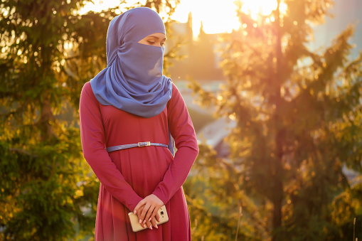 Portrait of beautiful Muslim woman with green eyes wearing blue scarf face closed covered with aveil background forest trees in the park.