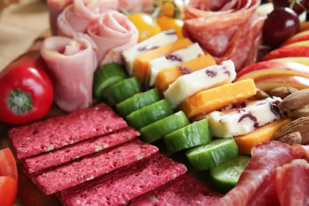 Stock photo showing close-up, elevated view of wooden charcuterie board covered with prepared sliced and chopped ingredients including rows of beetroot crackers, ham and salami roses, sliced cucumber, red apple, mini bell peppers, tomato flower-shape, Red Leicester, White Cheddar with cranberries, almonds and cherries.