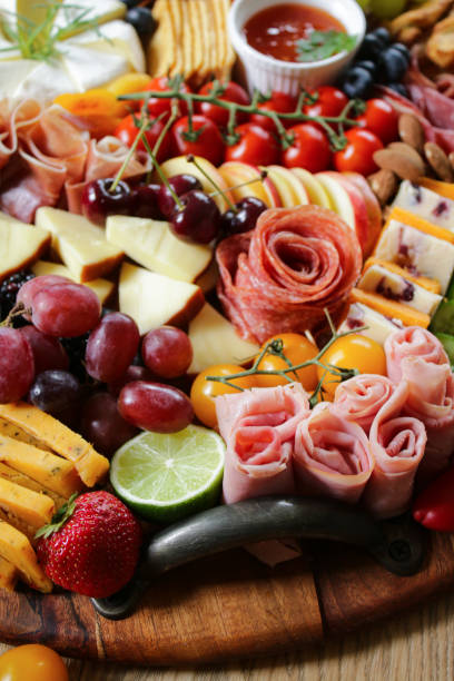 Stock photo showing close-up, elevated view of wooden charcuterie board covered with prepared sliced and chopped ingredients including rows of crackers, ham and salami roses, cherries, grapes, strawberries, blackberries, vine tomatoes, Smoked cheese, Brie, Red Leicester, Cheddar with cranberries, almonds, blueberries and a ramekin of chutney. imagen de tabla de charcutería, filas de galletas, rosas de jamón y salami, cerezas, uvas, fresas, moras, tomates de vid, queso ahumado, brie, red leicester, cheddar con arándanos, almendras, arándanos, ramekin de chutney, enfoque en primer plano