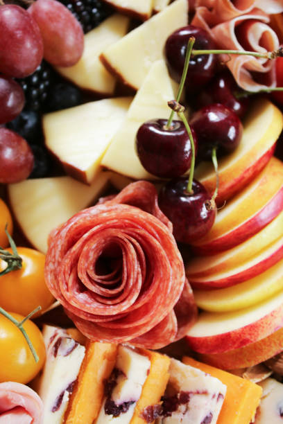 Full frame image of charcuterie board ingredients, rows of ham and salami roses, cherries, red grapes, yellow vine tomatoes, Smoked cheese, Red Leicester, Cheddar with cranberries, apple slices, blackberries and blueberries, elevated view