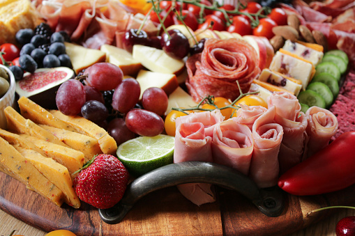 Stock photo showing close-up, elevated view of wooden charcuterie board covered with prepared sliced and chopped ingredients including rows of beetroot crackers, ham and salami roses, cherries, red grapes, strawberries, lime, mini red pepper, cucumber, fig  blackberries, vine tomatoes, Smoked cheese, Red Leicester, Cheddar with cranberries, almonds, blueberries and a ramekin of green olives.